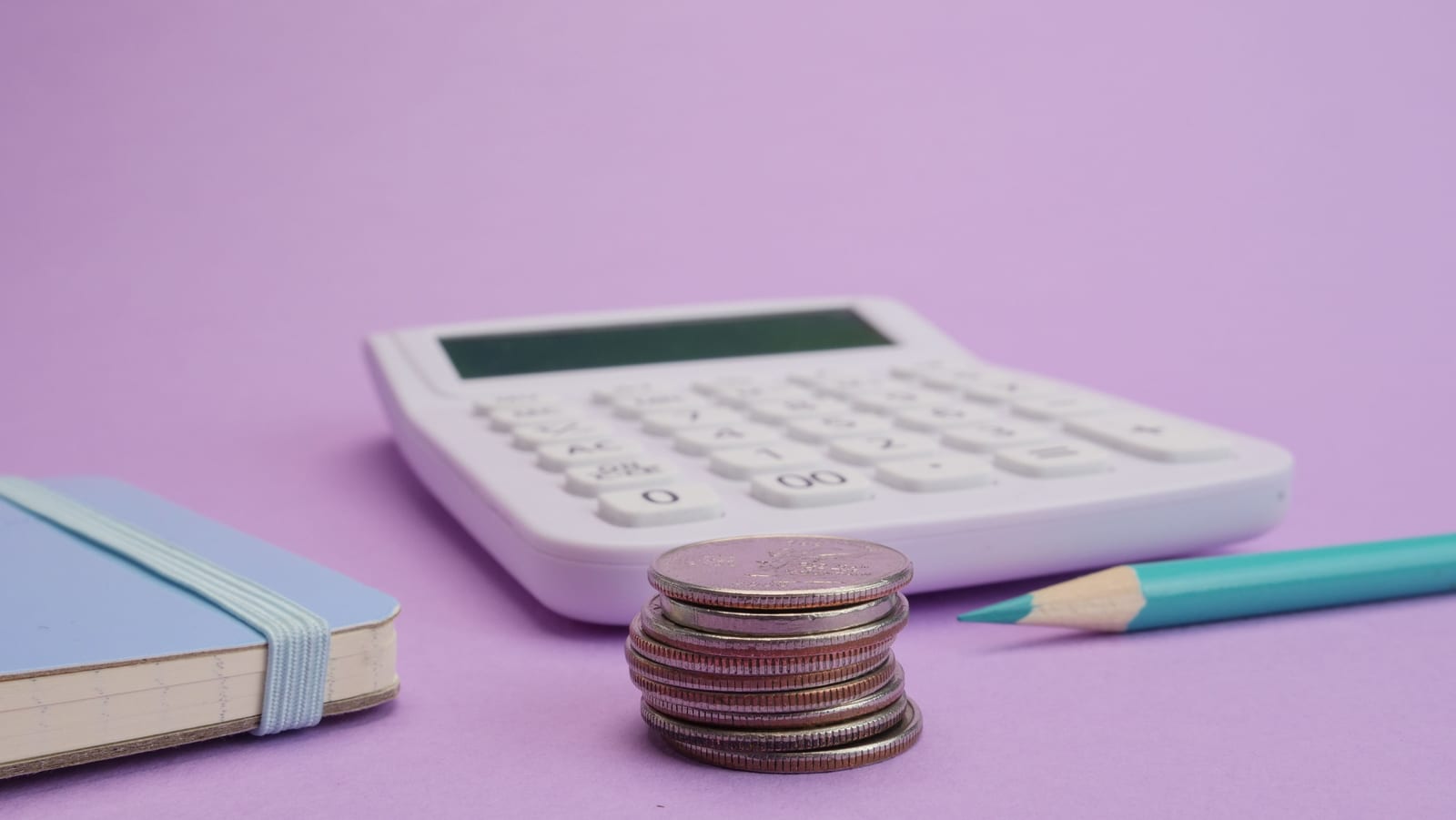 A white calculator, a stack of coins, a notebook and a pencil on a violet background
